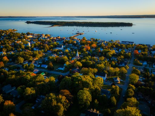 Aerial view of a coastal town with lush greenery and a serene bay at sunset, showcasing vibrant autumn colors and calm waters.