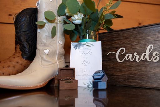 A wedding decor with cowboy boots, floral arrangement, invitation, and card box on a wooden table.