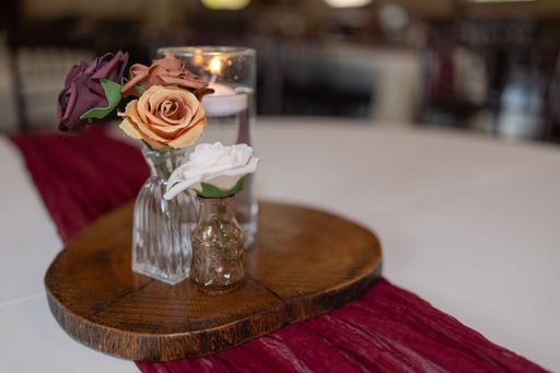An elegant table centerpiece with artificial roses in glass vases on a wooden base, featuring a candle and red fabric accent.