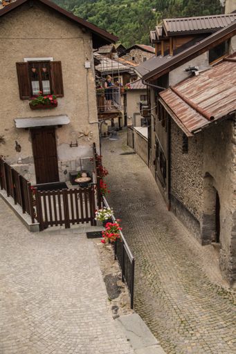 Charming cobblestone street in a quaint European village with rustic houses and vibrant flowers, featuring middle-aged individuals enjoying the view.