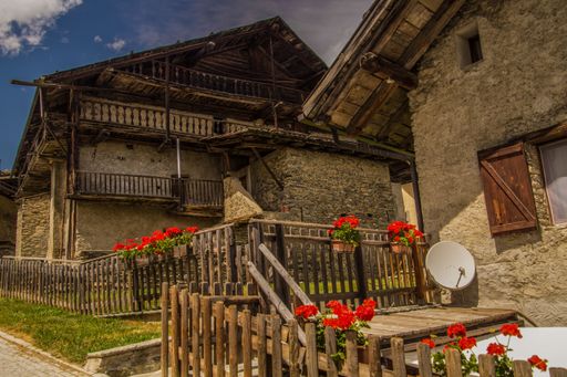 A rustic stone house with wooden balconies and red flowers under a blue sky in a quaint village setting.