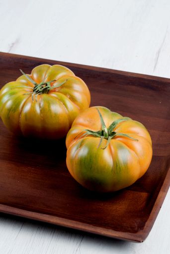 Two heirloom tomatoes on a wooden tray against a white background, showcasing their vibrant orange and green hues.