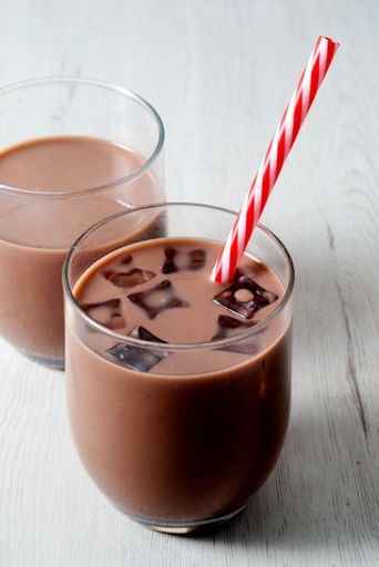 Two glasses of iced chocolate milk with a striped straw on a light wooden table.