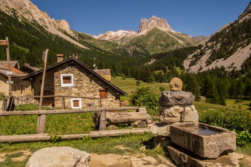 Charming alpine village scene with rustic stone house and mountain backdrop under clear blue sky in summer.