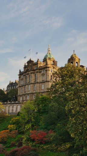 Historic building with Scottish flags and lush gardens in Edinburgh under a clear sky.