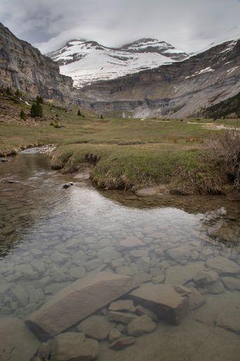 Scenic view of a serene mountain landscape with a clear stream and snow-capped peaks under a cloudy sky in Ordesa Valley, Spain.