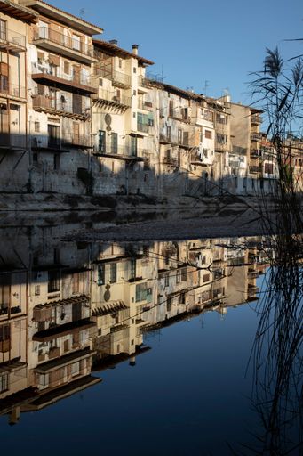 Charming riverside buildings reflecting in a calm river under a clear blue sky in Valderrobres, Spain.