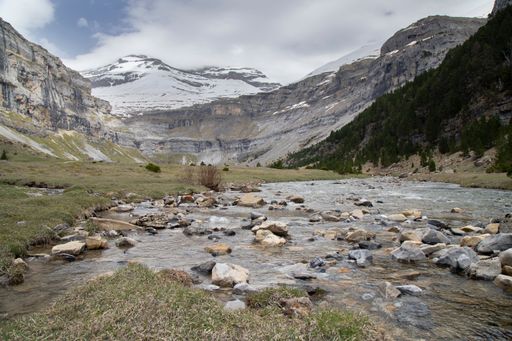 A serene mountain landscape with a flowing river and snow-capped peaks under a cloudy sky, captured in Ordesa Valley, Spain.
