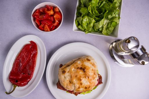 A top view of a burger with homemade bread and bacon with some tomatoes, lettuce and pepper near it