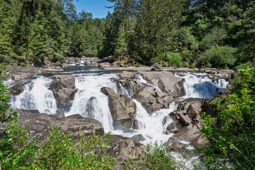 A scenic view of a cascading waterfall surrounded by lush greenery and rocks under a clear blue sky.