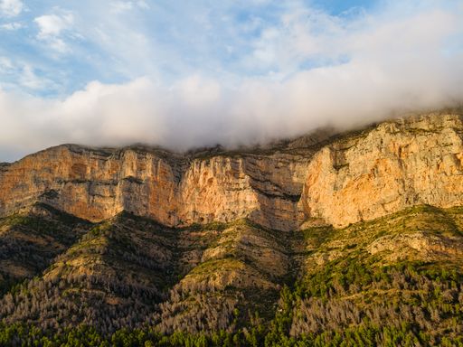 Montgo massif showing a mountain face with clouds covering its summit, with green and dry vegetation on the slopes.