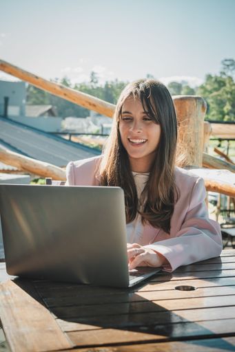 Young woman in casual attire working on a laptop outdoors, enjoying a sunny day at a wooden table.