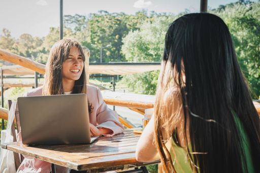 Two young women in a casual outdoor meeting, one with a laptop, enjoying a sunny day at a cafe with lush greenery in the background.