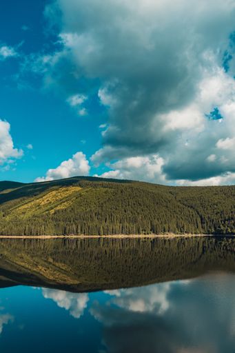 A vertical shot of forested mountains reflected in the water under a blue cloudy sky