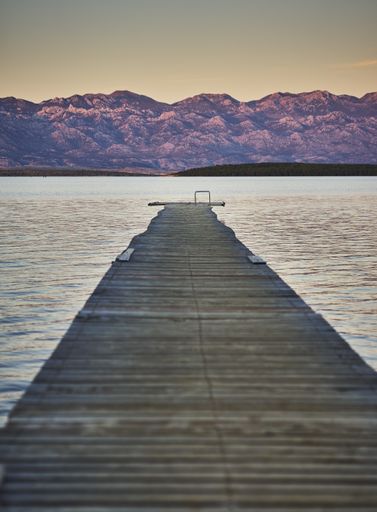 A vertical shot of a wooden walking path on a calm lake surrounded with hills captured on a sunny day