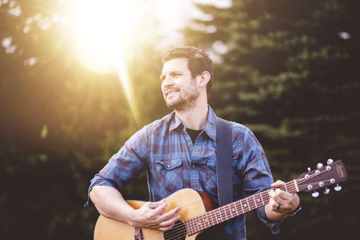 A young male in a park holding a guitar and playing a song from the Christian hymn book