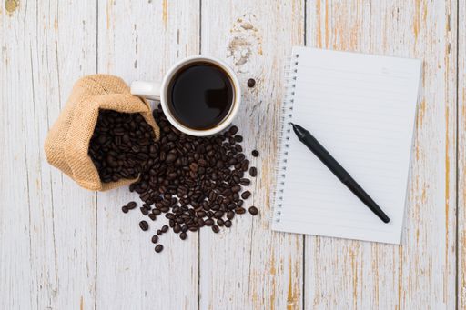 A top view of a cup of coffee and a sack of coffee beans with a notebook on a wooden desk