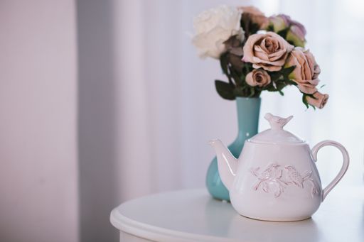 A selective closeup shot of a white ceramic teapot near blue vase with pink roses on a white table