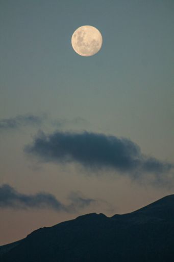 A vertical of a full moon illuminating the cloudy sky above the mountains in blue hour