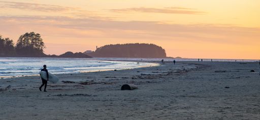 A sandy beach with a person holding a surfing board during the sunset