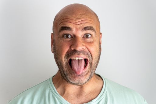 A close-up shot of a face of a bald bearded Caucasian man gesturing sticking out his tongue