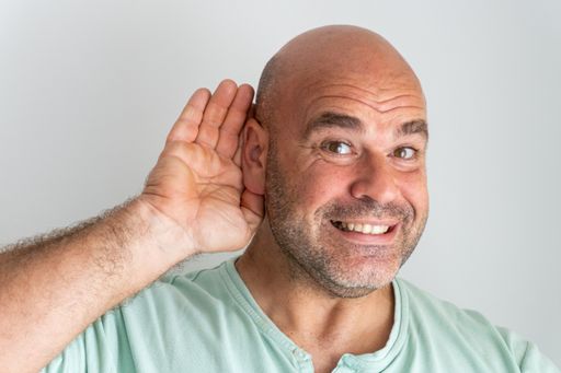 A close-up shot of the face of a bald and bearded Caucasian man gesturing with his hand behind his ear