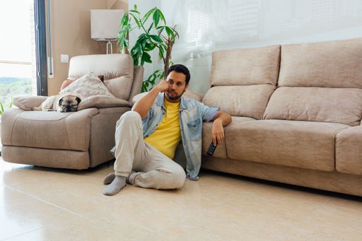 A horizontal shot of a male sitting on the floor and watching a TV with remote control in his hand