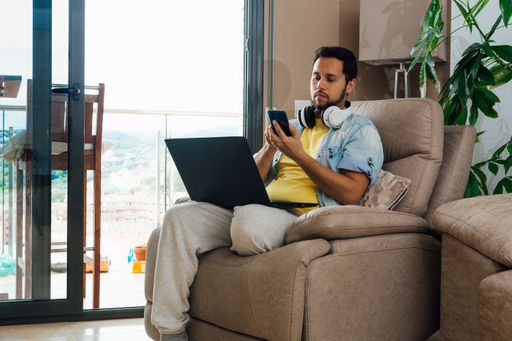 A young man with headphones checking his phone at home with a laptop on his lap