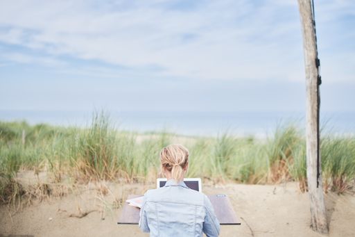 A back view of an unrecognizable blonde woman working on a laptop at a beach in summer