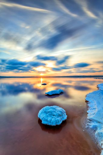 A vertical scenic view of a sunset reflection in the water with frosty stones under a cloudy winter sky in a long exposure - wallpaper