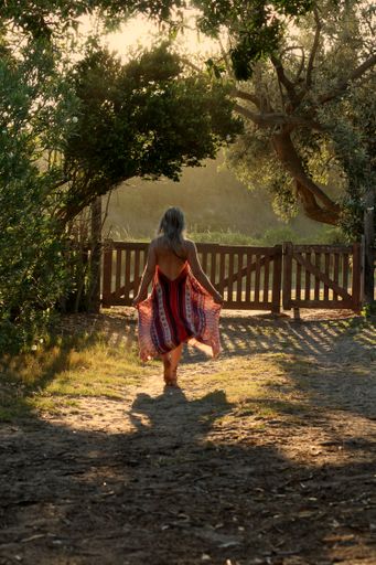 A scenic shot of a woman wearing a dress from her back walking to the fence in the forest