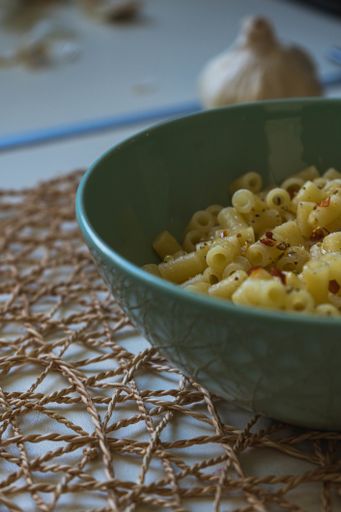 A soft focus of cooked seasoned rigatoni pasta in a green bowl on a burlap mesh with a garlic bulb in the background