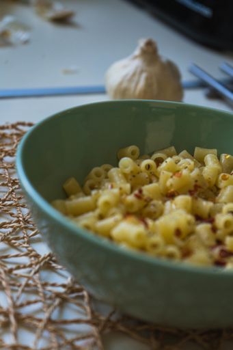 A soft focus of cooked seasoned rigatoni pasta in a green bowl on a burlap mesh with a garlic bulb in the background