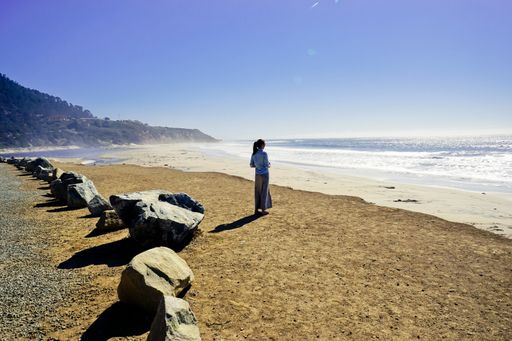 A female standing on a shore near rocks and the water with mountain and clear blue sky in the background