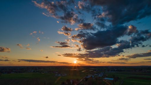 An aerial shot of a field and houses with a beautiful sunset and a cloudy sky