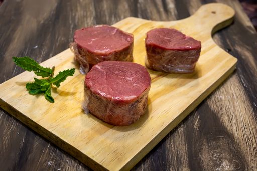 A closeup of three pieces of fresh raw beef meat wrapped in plastic bags on a cutting board