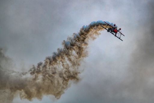 A low-angle shot of a biplane in a flight against a gray sky with a trailing smoke during the airshow