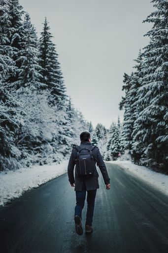 A vertical shot of a young man with a backpack walking down the road in a forest covered in the snow