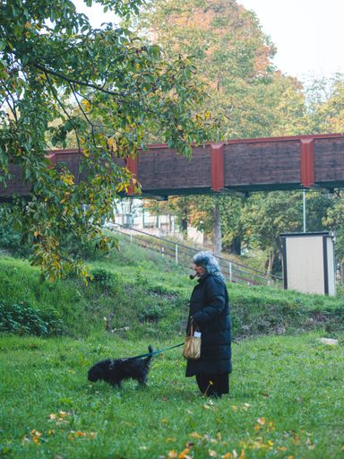 A vertical shot of an older female in a black coat smoking a pipe while walking with her dog in a park