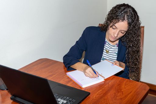 Young woman working from home with a laptop computer while taking notes in a notebook.