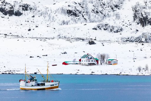 Fishing boat sailing through a fjord in Lofoten, Northern Norway, with snow on the mountains. There's an old farm in the background.