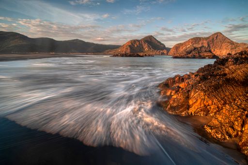 Late afternoon sun illuminates Partatutai Island rocks and black volcanic sand of Whatipu Beach