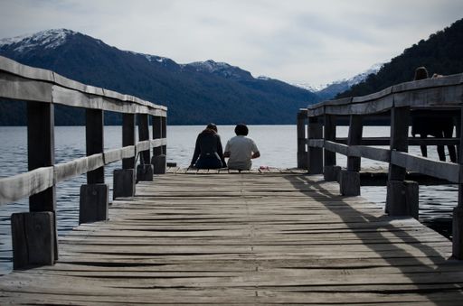 Two people sitting and chatting on a pier with a lake and mountains in the background