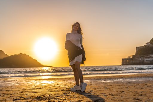 A young Colombian blonde female in a grey dress and a pullover walking at the beach of San Sebastian, Spain at sunset