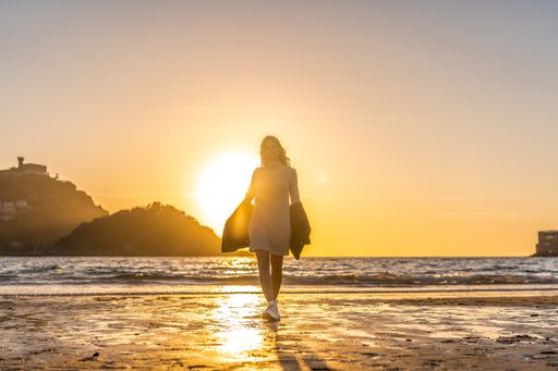 A young Colombian blonde female in a grey dress and a pullover walking at the beach of San Sebastian, Spain at sunset