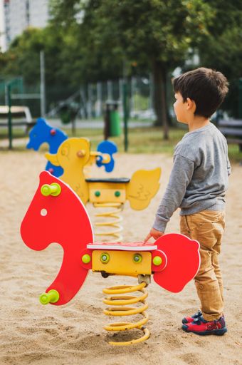 A vertical shot of a young Caucasian boy standing next to a rocking chair in a playground