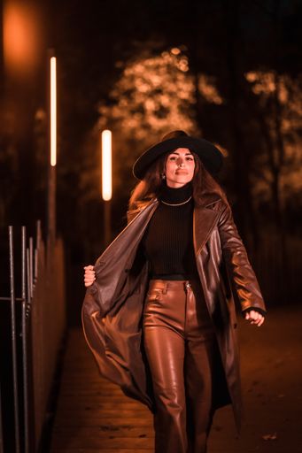 A vertical shot of a Caucasian female posing in a hat at night in a raincoat on a parcel, illuminated with dim orange light from the side