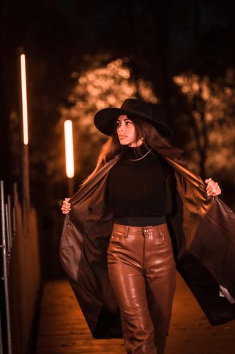 A vertical shot of a Caucasian female posing in a hat at night in a raincoat on a parcel, illuminated with dim orange light from the side