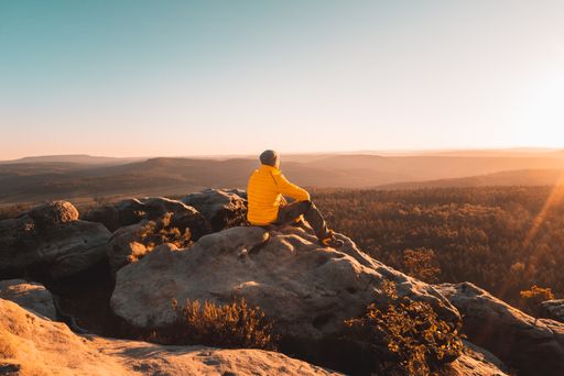 A back view of a young male hiker sitting on a huge rock admiring the view of nature on a sunny day
