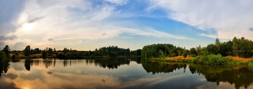 A panoramic shot of a tranquil lake with trees reflected on it under a cloudy sky during sunset